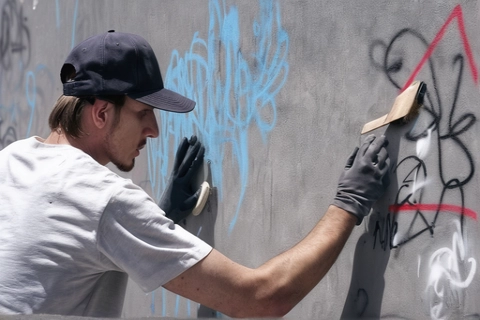 Mann in weißem T-Shirt und Kappe entfernt Graffiti von einer Wand mit einem Handwerkzeug und trägt dabei Schutzhandschuhe.